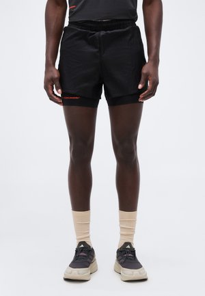 Homme portant un short de course noir superposé, des chaussettes beiges montantes et des baskets Adidas noires, debout sur un fond blanc uni.
