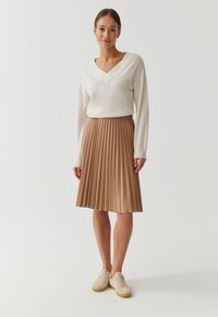 Beige geplooide rok van zachte stof, gecombineerd met een witte V-halstrui en beige sneakers. Gladde textuur met duidelijke plooien.