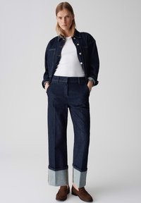 Femme portant une veste en jean foncé et un jean retroussé avec des mocassins marron et un haut blanc, se tenant contre un fond gris uni.