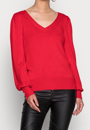 Rot gestrickter Pullover mit V-Ausschnitt, langen Puffärmeln und gerippten Manschetten, kombiniert mit schwarzen Kunstlederhosen. Akzentuiert mit kleinen Hardware-Details.