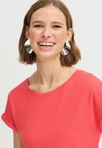 Korallenfarbenes T-Shirt mit kurzen Ärmeln und rundem Halsausschnitt. Auffällige glänzende silberne abstrakte Ohrringe. Glatte Textur, lässiges Design.