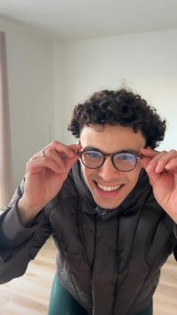 Bruine, ronde bril die wordt vastgehouden door een persoon met krullend haar, gekleed in een donker donzen jasje. Op de achtergrond zijn lichtgekleurde muren en een houten vloer te zien.