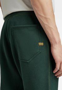 Pantalones de chándal verde oscuro con un bolsillo trasero, costura en contraste y una etiqueta de marca. El material parece suave y texturizado.