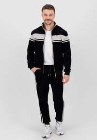 Chándal de terciopelo negro con rayas grises, chaqueta con cremallera, puños acanalados y pantalones a juego con cordón, con acentos en oro en el logo. Camiseta blanca y zapatillas.