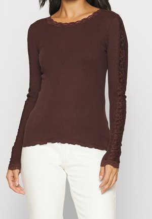 Femme portant un haut marron ajusté à manches longues avec des détails en dentelle sur les manches et des bords festonnés, associé à un pantalon blanc.