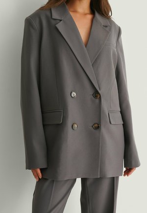 Vrouw die een oversized donkergrijze double-breasted blazer met bijpassende broek draagt, staand tegen een effen lichte achtergrond.