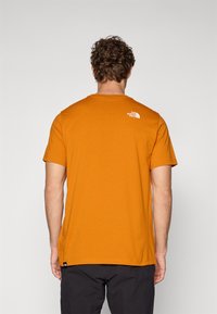 Tricou portocalie cu mânecă scurtă, din bumbac, cu decolteu rotund și un mic logo alb pe spatele superior. Design simplu, culoare solidă.