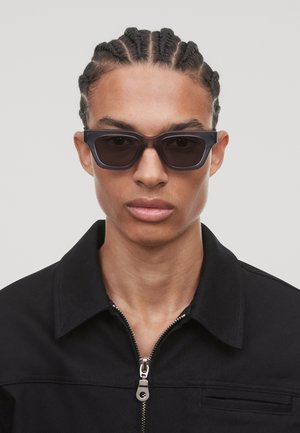 Des lunettes de soleil avec une monture rectangulaire noire et des verres teintés. Portées par un mannequin avec des cheveux tressés, associées à une veste noire à fermeture éclair.