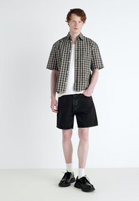 Kortærmet plaid skjorte i grøn og beige over en hvid t-shirt, parret med sorte shorts og sorte sko. Hvide sokker synlige.