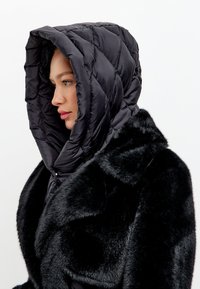 Zwarte gewatteerde pufferjas met een capuchon, featuring een gestikt ontwerp en nepbont accenten op de schouders, benadrukt warmte en comfort.