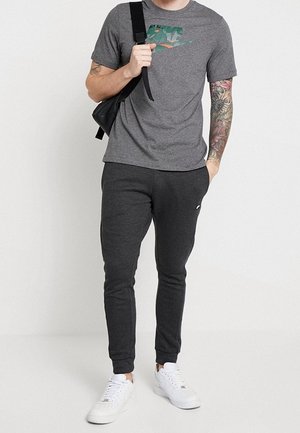 Šedé tričko s krátkým rukávem se zeleno-oranžovým vzorovaným logem, doplněné tmavě šedými zúženými tepláky a bílými teniskami. Černá kabelka přes rameno.