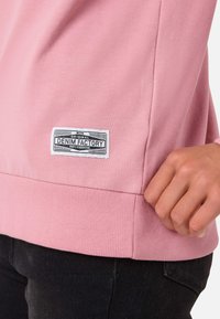 Ljusrosa sweatshirt i bomullsblandning, med ribbat avslut och en vävd etikett med "Denim Factory"-logotyp på sidan.