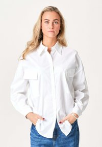 Blond kvinde med løst hår iført en hvid skjorte med knapper og forlommer samt blå jeans, hænderne i lommerne, neutral baggrund.