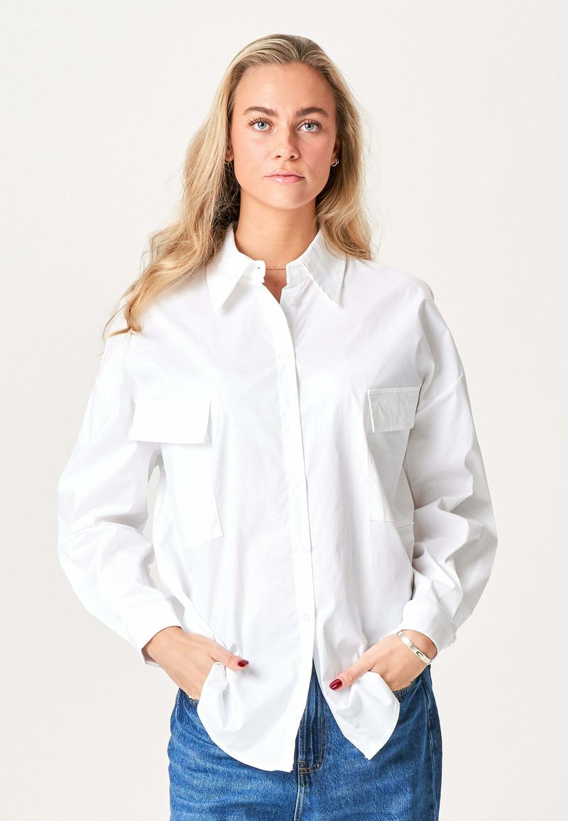 Blond kvinde med løst hår iført en hvid skjorte med knapper og forlommer samt blå jeans, hænderne i lommerne, neutral baggrund.