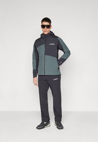 Herre Terrex-jakke i mørkegrå og teal, med glidelås foran, hette og reflekterende detaljer; kombinert med svarte bukser og grå sneakers.