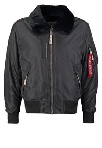 Veste bomber noire avec col en fourrure, fermeture éclair à l'avant, poignets et ourlet côtelés, poches latérales et une étiquette en tissu rouge sur le bras gauche.