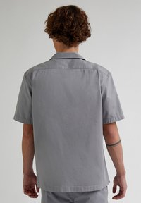 Camicia a maniche corte in tessuto grigio chiaro, caratterizzata da un colletto a camicia, cuciture impunturate e un orlo dritto con spacchetti laterali.