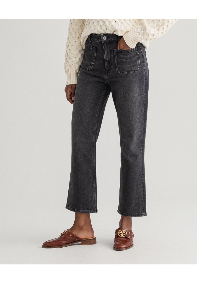 Personne portant un jean noir taille haute raccourci, un pull crème texturé et des mocassins en cuir marron à enfiler avec un détail de chaîne dorée.