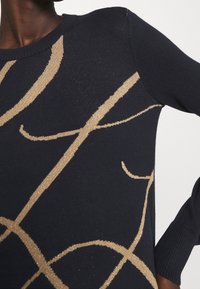 Marinblå stickad tröja med guldmetallictråd i abstrakta mönster, rund halsringning och ribbade ärmslut, som visar en mjuk textur.