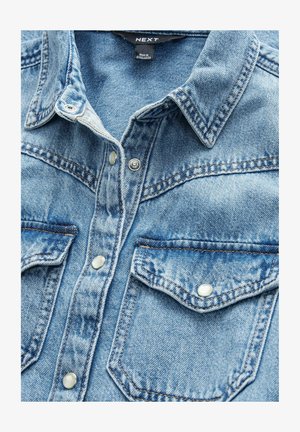 Camisa de mezclilla de color azul claro con dos bolsillos frontales, botones a presión y cuello puntiagudo. Tejido texturizado con detalles de costura visibles.