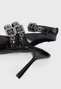 Juodas aukštakulnis sandalai su blizgia oda, sagtimis ir inkarais. Atvira konstrukcija pasižymi elegantiška stiletto kulniuku.