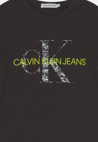 Černé bavlněné tričko s velkým stříbrným nápisem "ck" a "CALVIN KLEIN JEANS" v živé limetkové zelené barvě pod ním. Hladká textura.