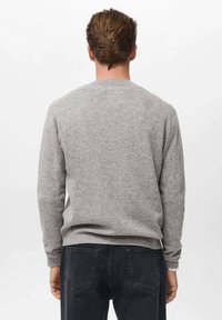 Suéter gris de punto con puños y dobladillo de canalé, con un escote redondeado y una textura suave. Lleva puesto sobre unos jeans oscuros.