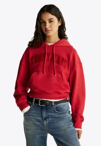 Felpa rossa con la scritta "TOMMY JEANS New York" stampata in rosso scuro. Presenta una tasca frontale, cappuccio con stringhe e maniche ampie, abbinata a jeans blu.