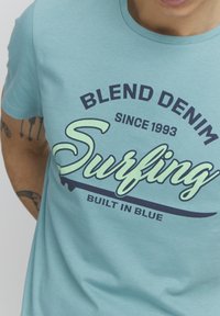 Gaišzils T-krekls no mīksta auduma, ar lielu grafiku, uz kuras redzams teksts "Blend Denim" un "Surfing" tumši zilā un gaiši zaļā krāsā.
