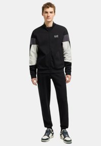 Tuta nera con accenti grigi e bianchi, caratterizzata da una giacca con zip e pantaloni abbinati, realizzata in un materiale liscio ed elastico. Logo EA7.