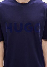 Tamsiai mėlynas medvilninis marškinėlis, papuoštas ryškiu, blizgiu "HUGO" logotipu šiek tiek tamsesnėje mėlynoje spalvoje. Klasikinis apvalus kaklas ir trumpomis rankovėmis.
