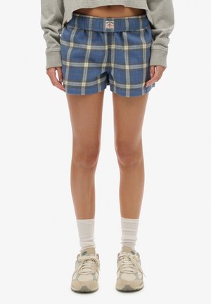 Person iført blå og hvide ternede shorts, hvide sokker, beige sneakers og en grå langærmet cropped top står.