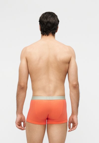 Mann mit dunklem Haar steht mit dem Rücken zur Kamera, trägt orangefarbene Boxershorts mit grauem Bund vor einem einfachen hellen Hintergrund.