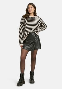 Frau in einem schwarz-beige gestreiften Pullover, schwarzer Lederrock, transparenter Strumpfhose und schwarzen Schnürstiefeln, die mit einer Hand in die Hüfte gestützt dasteht.