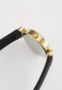Montre dorée avec un boîtier rond, un cadran argenté et un bracelet en silicone texturé noir. Comprend une couronne sur le côté pour régler l'heure.