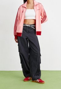 Donna con un giubbotto a track rosa Adidas, un crop top bianco, pantaloni cargo neri larghi e sneakers rosse Adidas in piedi su un pavimento verde.