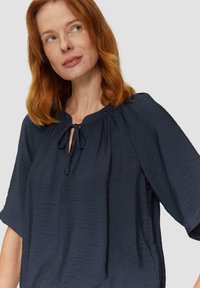 Granatowa bluzka o teksturowanym wykończeniu, z okrągłym dekoltem, detalem w postaci wiązania oraz szerokimi rękawami. Lekka, casualowa konstrukcja zapewniająca komfort.