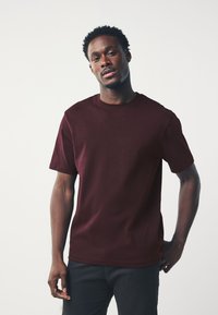 Camiseta burdeos de manga corta en tejido de algodón suave, con diseño de cuello redondo y aberturas laterales. La modelo la lleva con pantalones negros, posando frente a un fondo claro.