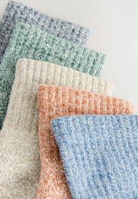 Cinq paires de chaussettes en maille texturée, de couleur bleue, orange, beige, verte et violette, sont soigneusement empilées sur une surface claire.