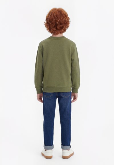 Παιδί με κόκκινα σγουρά μαλλιά, φοράει ελιάς πράσινο φούτερ, μπλε τζιν με μανσέτες και λευκά αθλητικά παπούτσια, στέκεται γυρισμένο με την πλάτη σε λευκό φόντο.