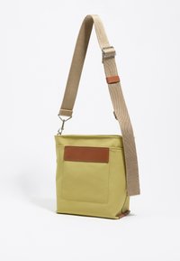 Borsa sintetica gialla con un accento in pelle marrone, forma rettangolare e tracolla regolabile in tela beige. Presenta una tasca frontale senza chiusura.