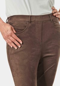 Bruine suède broek met een gladde textuur, met voorzakken en een getailleerd ontwerp, weergegeven op heuphoogte met een hand erop steunen.