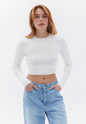 Gekürztes weißes, geripptes Langarmoberteil, kombiniert mit lockeren hellblauen Jeans. Das Model steht mit den Händen in den Taschen vor einem schlichten Hintergrund.