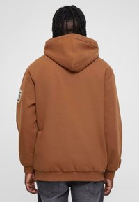Felpa con cappuccio marrone realizzata in tessuto morbido, caratterizzata da una vestibilità comoda, polsini a costine e un logo ricamato sulla spalla sinistra.