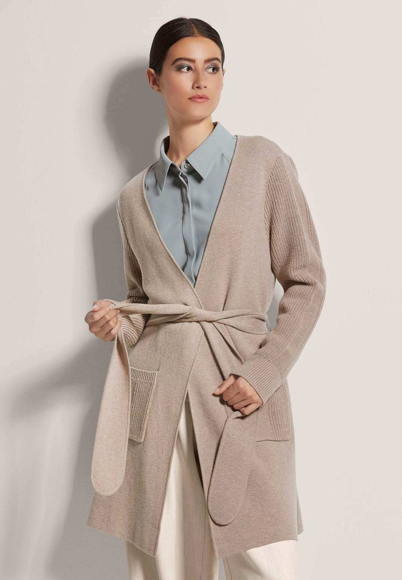 Beige Strickjacke mit gebundenem Taillenzug, offener Front und gerippten Ärmel Details. Getragen über einem hellblauen Hemd mit Kragen, kombiniert mit hellen Hosen.