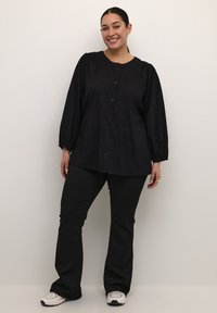 Blusa nera a maniche lunghe con spalle a sbuffo, apertura con bottoni e texture a pois, abbinata a pantaloni neri e sneakers con dettagli argentati.