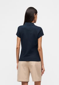 Μπλε μαρινιέρα πόλο με κλασικό γιακά, κοντά μανίκια και εφαρμοστή γραμμή, συνδυασμένη με μπεζ σορτς, που παρουσιάζει μια ομαλή υφή βαμβακιού.