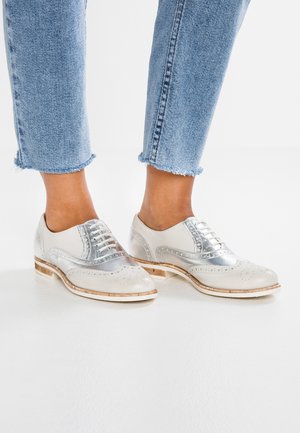 Füße, die hellblaue kurz geschnittene Jeans und weiße Leder-Brogue-Schuhe mit silbernen Details und Holzsohlen auf einem weißen Boden tragen.