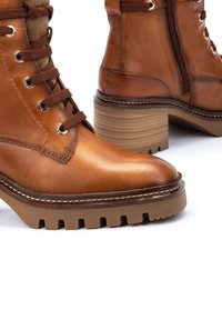 Botas de tobillo de cuero marrón con suela robusta y estriada, costuras decorativas, ojales dorados y acentos texturizados en la parte superior.