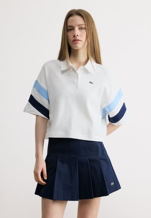 Noor naine, kes kannab valget polo-särki siniste triipudega ja tumesinist volditud seelikut, seisab ühetaolise heleda tausta ees.